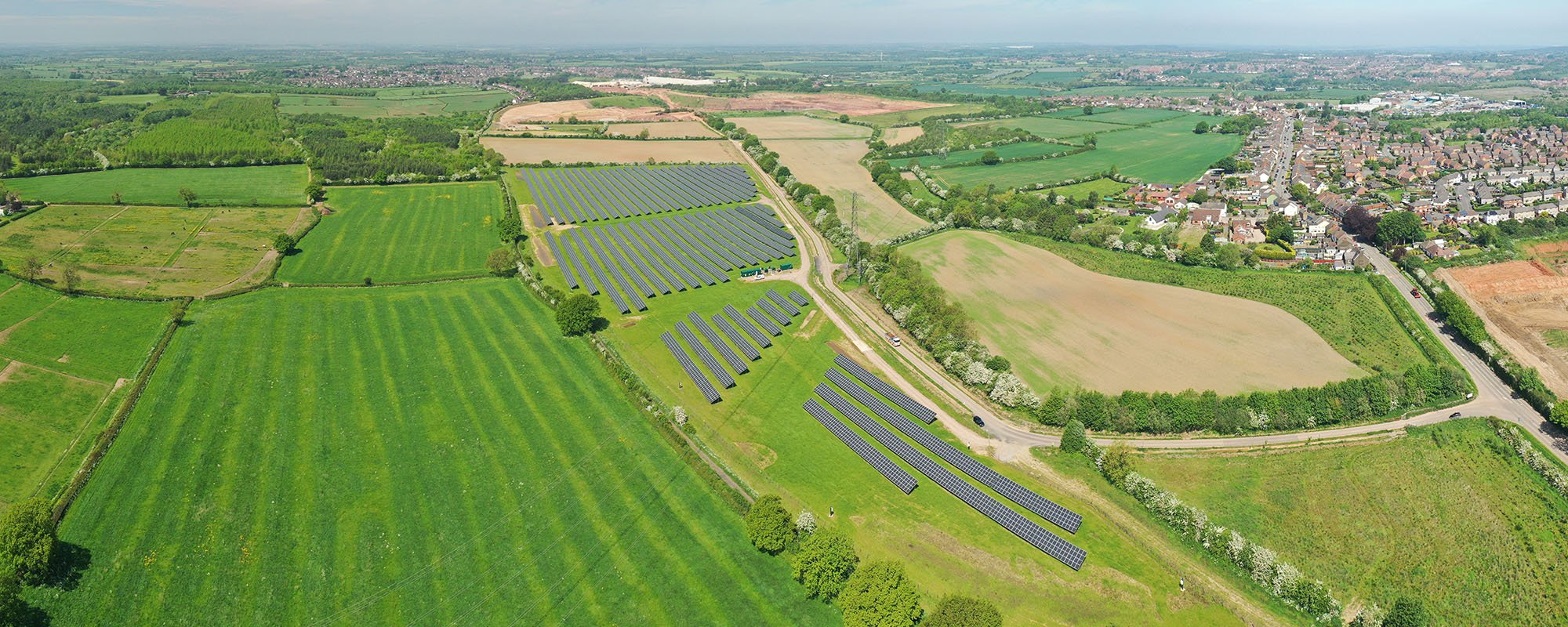 Aerial view of the Lightsource bp lbstock solar project in the UK, part of their UK Projects portfolio, rows of panels installed across vibrant green fields near a residential area under a bright sky.