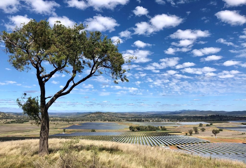 Rural landscape view from a hill, showing a large, sprawling utility-scale solar farm under a bright blue sky with wispy clouds