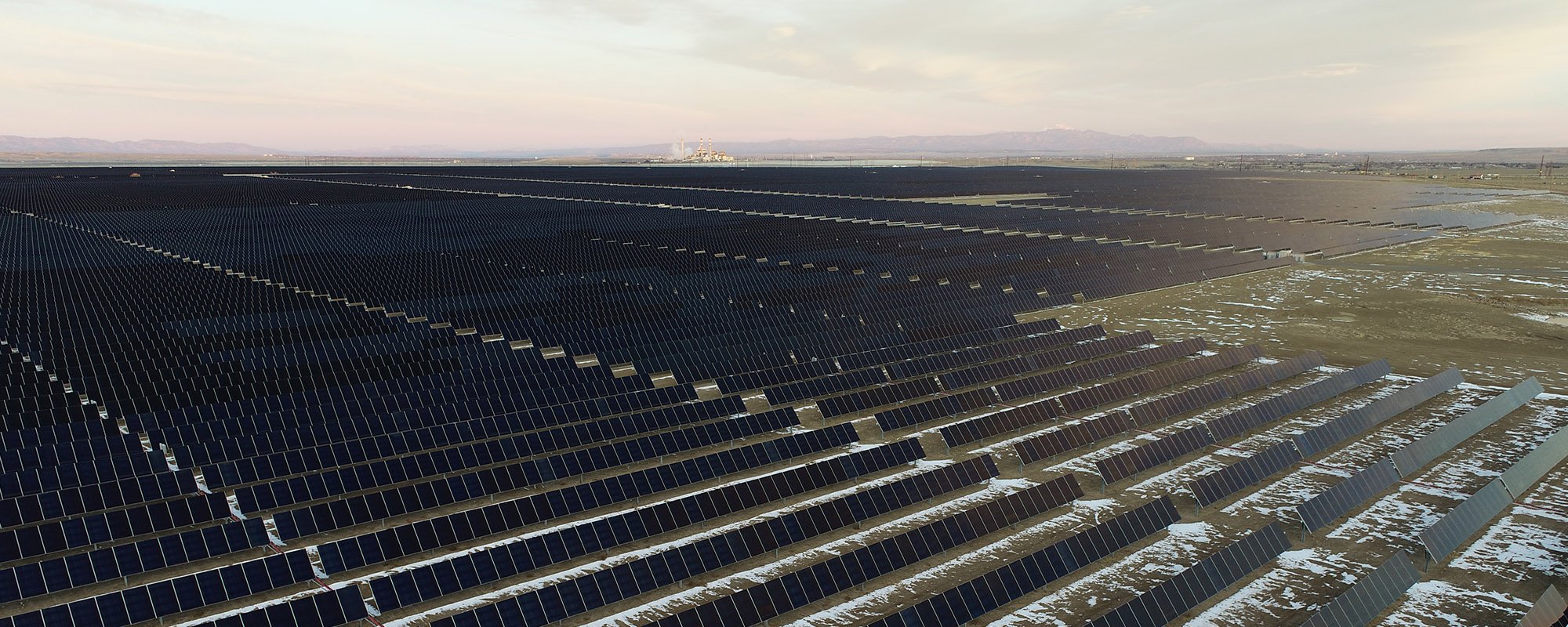 Aerial view of the Lightsource bp Sun Mountain Solar Farm in Colorado, part of their US Projects portfolio, vast rows of panels stretch across snow-dusted fields under a soft twilight sky, with distant mountains and industrial structures on the horizon.