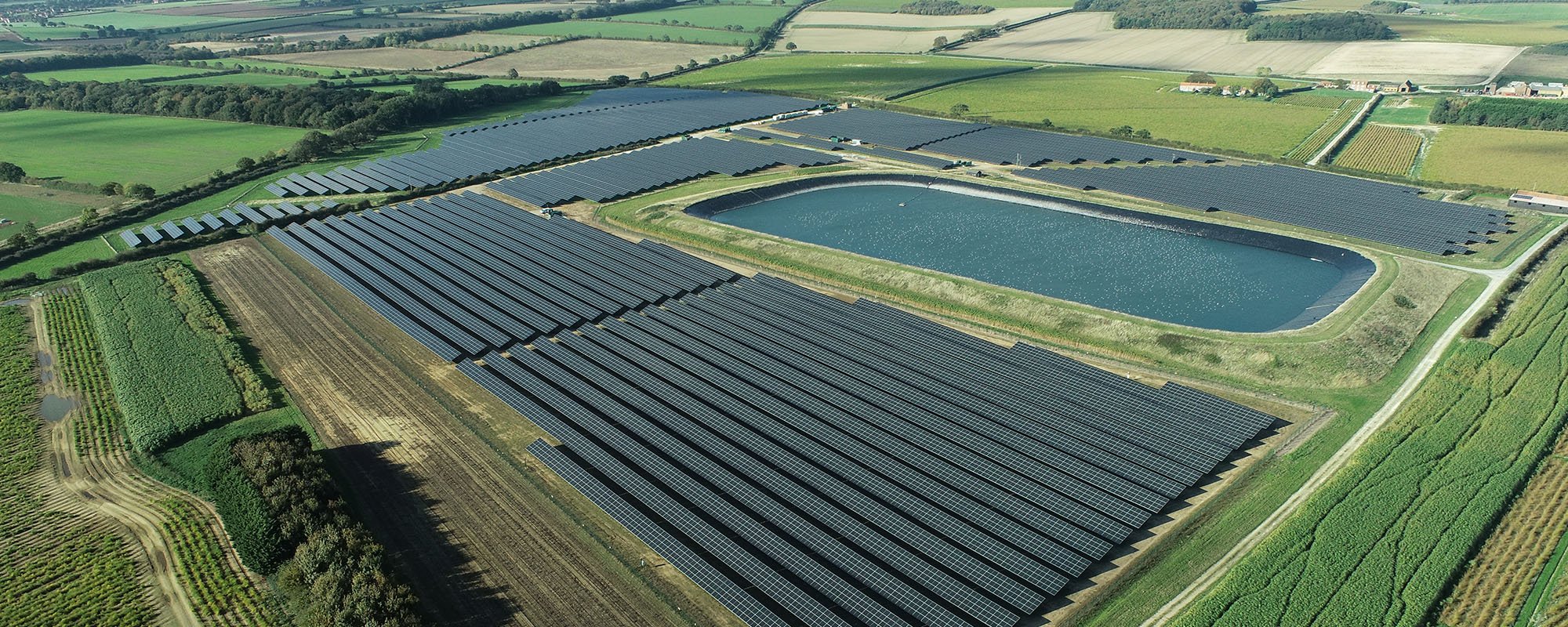 Aerial view of the Lightsource bp Thornham solar plant near Hunstanton, Norfolk — part of their UK Projects portfolio, with vast rows of panels installed across green fields surrounding a large reservoir.