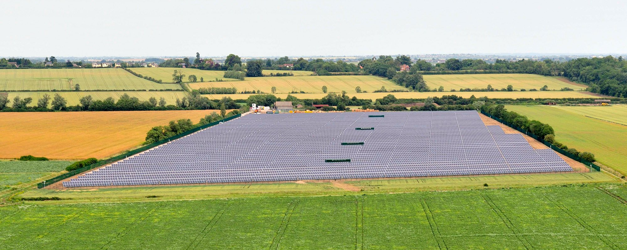 Aerial view of the Lightsource bp Wilburton solar farm in the UK, part of their UK Projects portfolio, a vast rectangular array of panels installed across green fields, bordered by trees and adjacent to golden farmland.