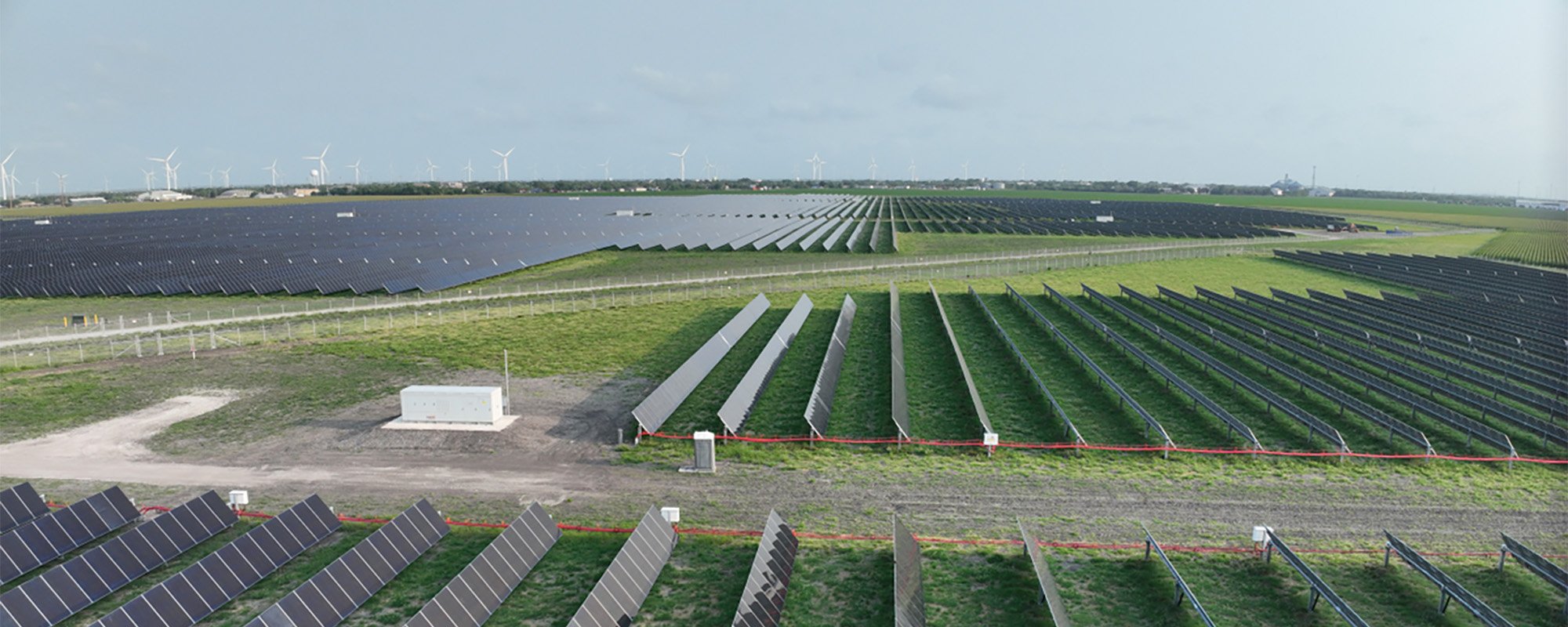 Aerial view of the Lightsource bp Peacock solar project in Texas, part of their US Projects portfolio, vast rows of solar panels stretch across green fields with wind turbines visible on the horizon.