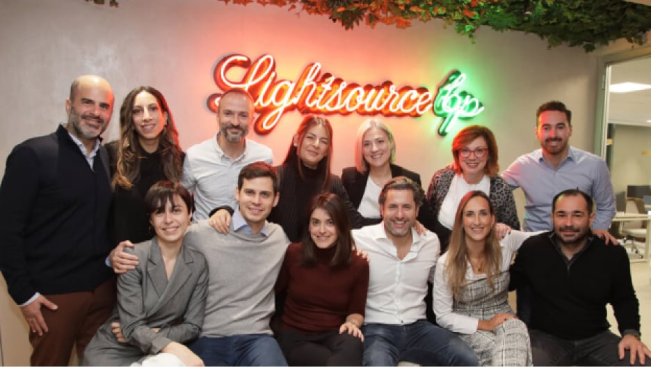 A group of thirteen people posing together indoors, smiling in front of a neon 
