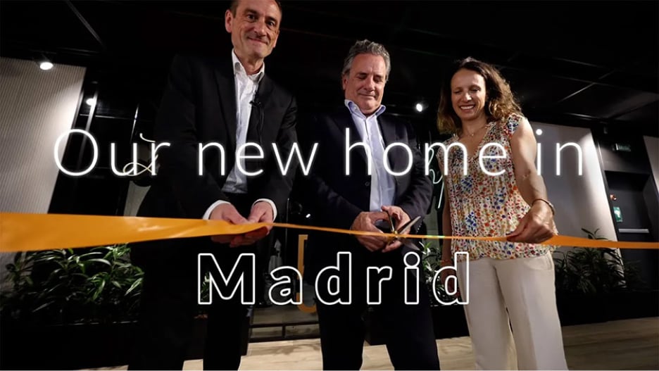 Three people stand cutting an orange ribbon under the text, 