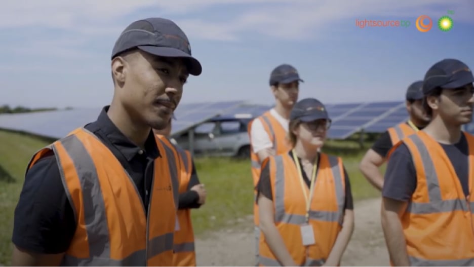 Group of people in orange safety vests and caps stand near solar panels on a solar farm.