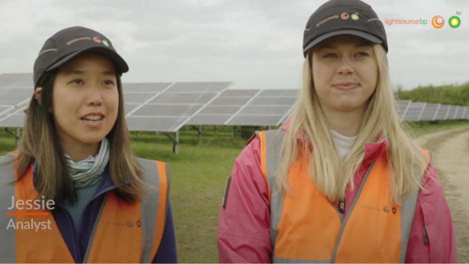 Two people in orange safety vests and caps stand in front of solar panels on a solar farm.
