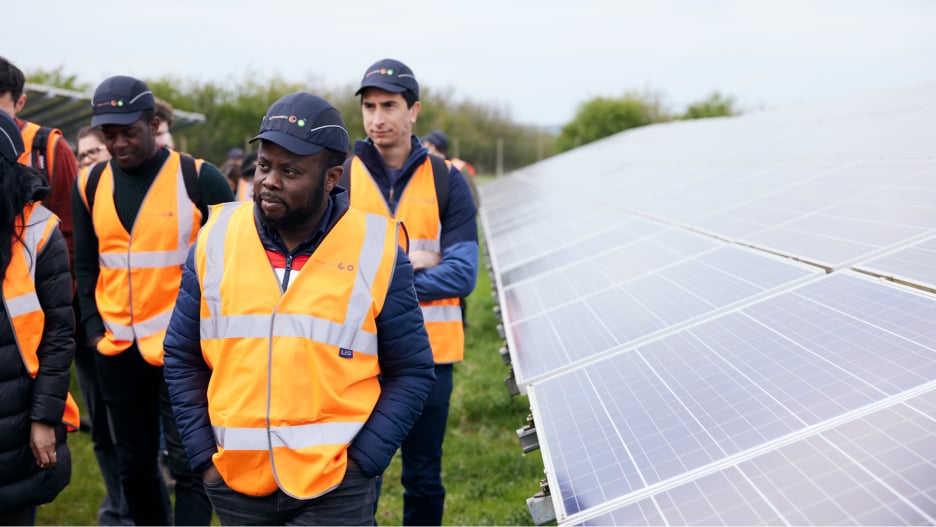 Group of people in orange safety vests and caps stand near solar panels on a solar farm.
