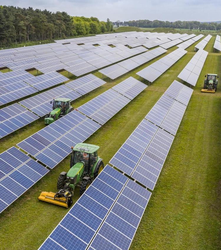 Wide aerial view of solar panels spanning a large grassy area, with tractors working between rows of panels.