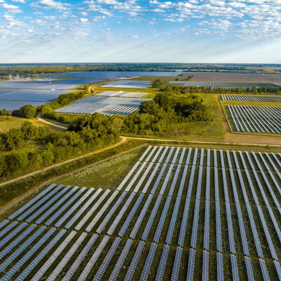 An aerial view of a large solar farm with rows of solar panels, surrounded by green fields and a body of water