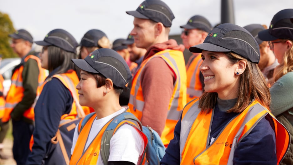 A group of people in orange safety vests and black caps are standing outdoors, attentively facing forward.