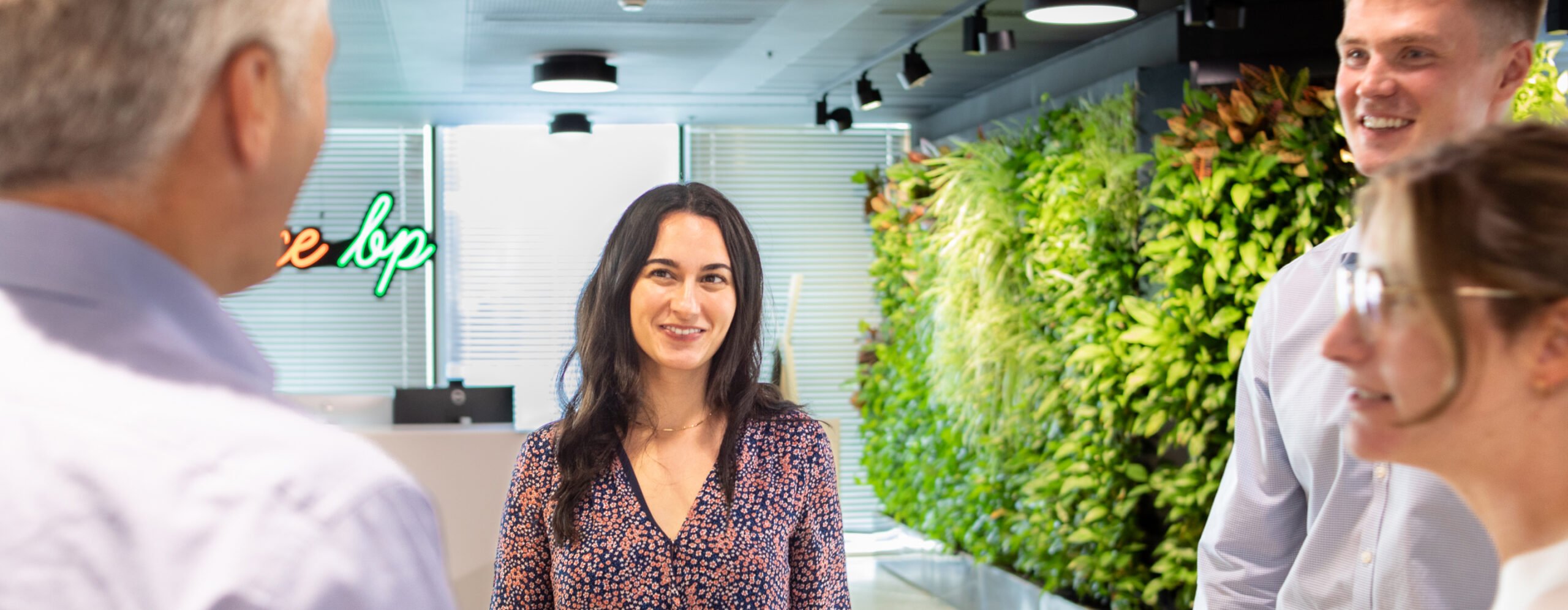 Four people are standing and talking in an office with a vertical garden wall and a neon sign in the background.