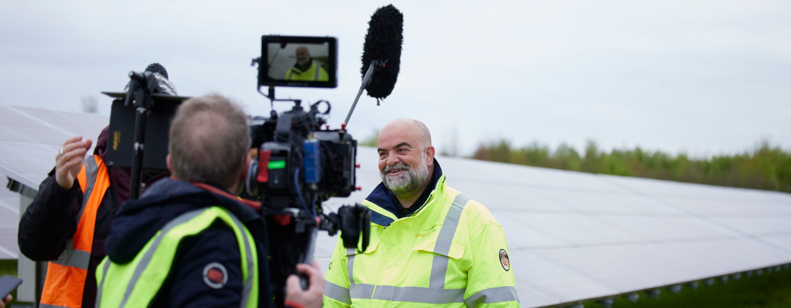 A man in a high-visibility jacket smiles while being filmed in front of solar panels, with a cameraman in focus.
