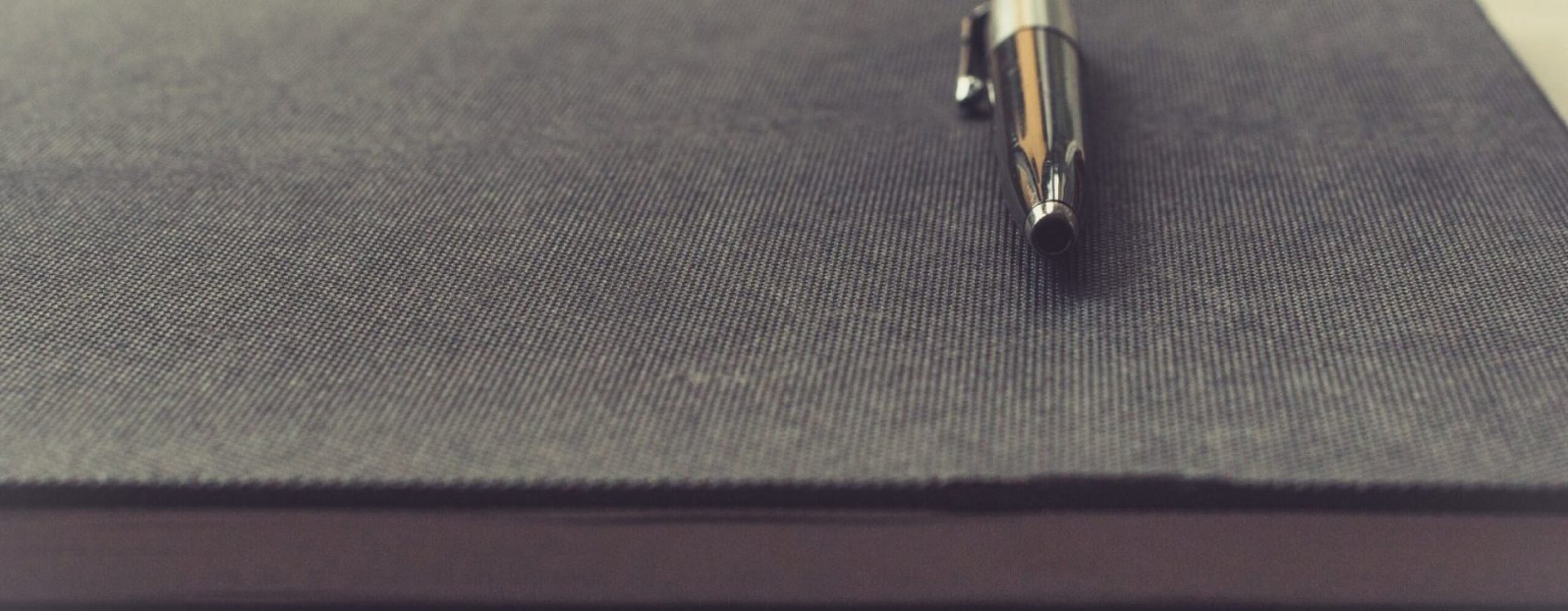 A close-up image of a silver pen resting on a dark notebook, suggesting a moment of contemplation or preparation for writing.