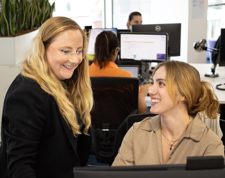 Two women smiling and talking at a desk in an office setting, with computers and people working in the background.