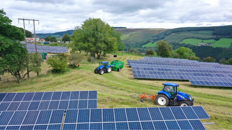 Photo showcasing agrisolar practices, with tractors mowing grass between rows of solar panels, demonstrating a sustainable and multi-functional use of land.