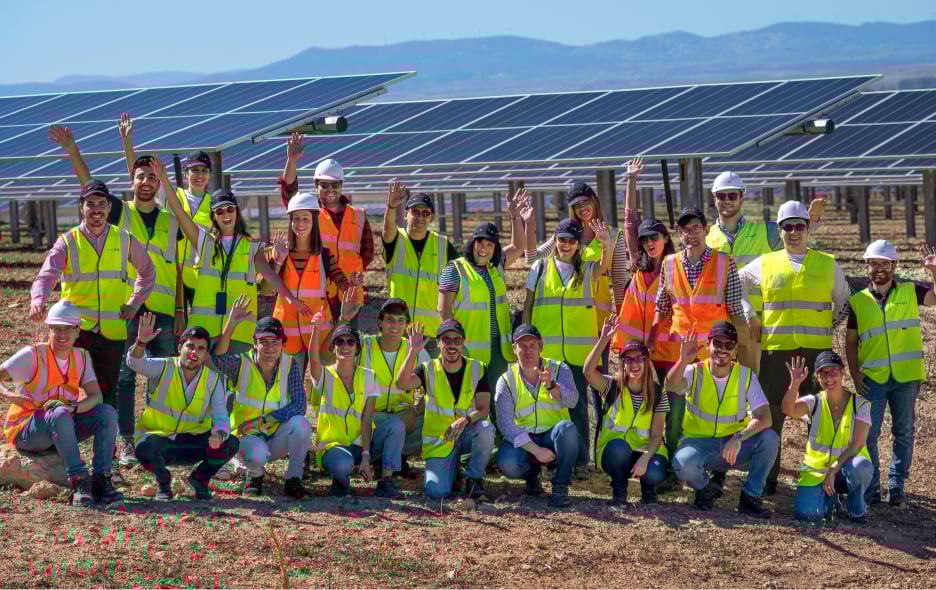 Group of people in safety vests and helmets pose in front of a solar panel array in a sunny landscape.