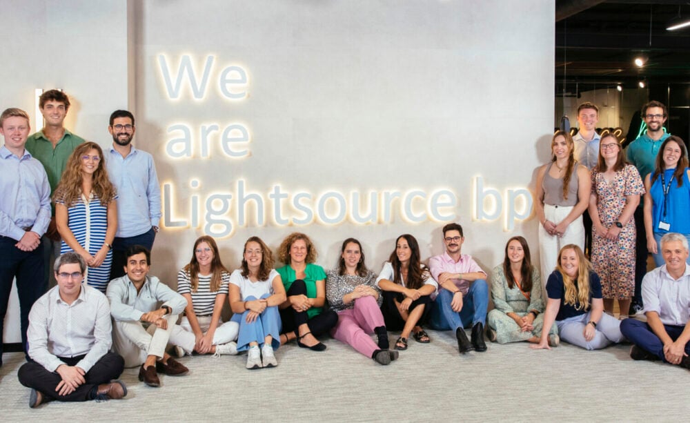A group of 19 people sitting and standing in front of a wall with the text 