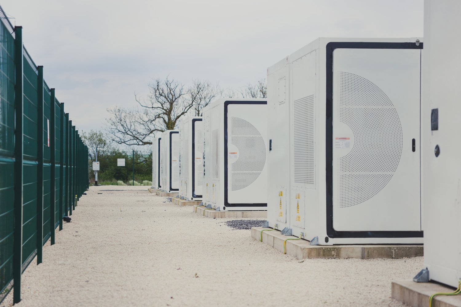 Row of white, industrial-scale Battery Energy Storage System (BESS) units, enclosed within a green perimeter fence at a utility-scale solar farm site, demonstrating energy storage infrastructure and security.