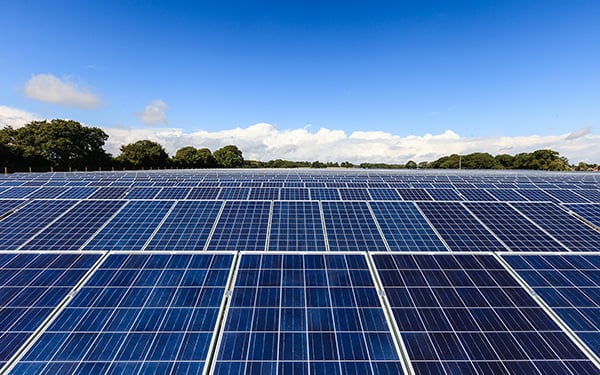 Rows of solar panels at a Lightsource bp solar project, installed under a bright blue sky with fluffy white clouds and a tree line on the horizon.