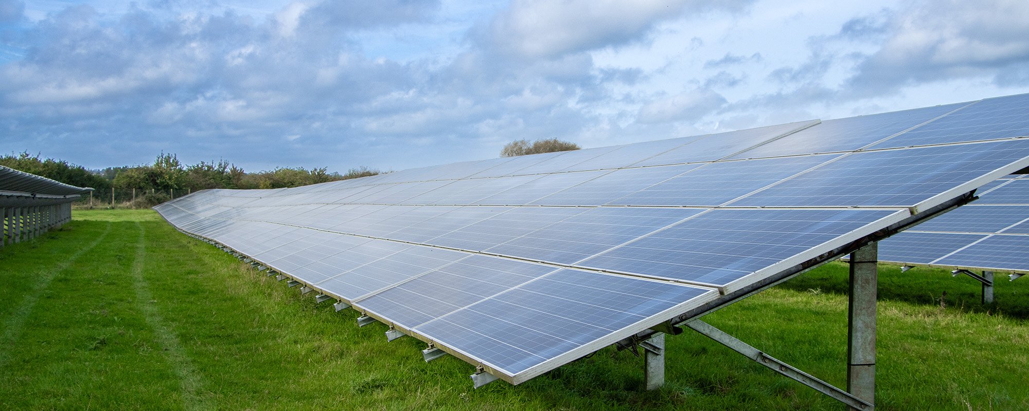 Rows of solar panels at a Lightsource bp solar project, installed across green grass under a partly cloudy blue sky.