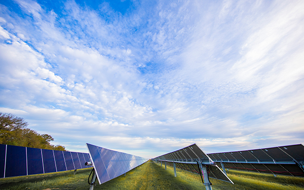 Rows of solar panels at a Lightsource bp solar project, stretching across green grass under a vast blue sky with scattered white clouds.