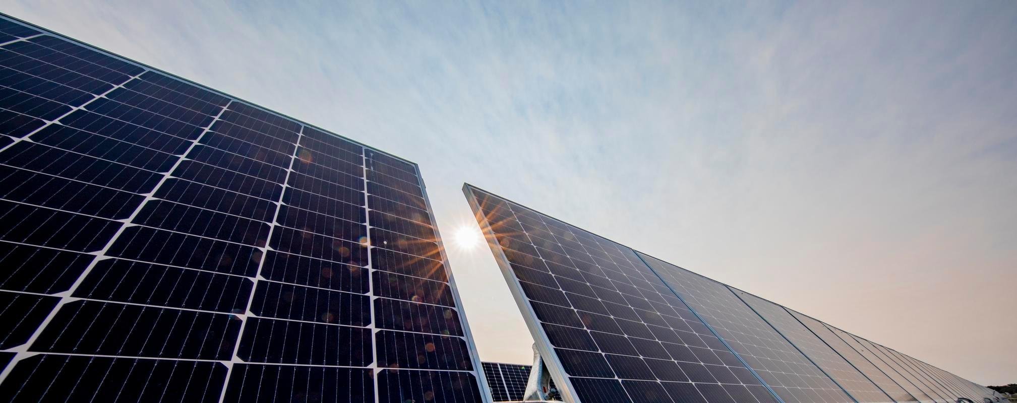 Close up, low angle shot of solar panels against a blue sky with light clouds, the sun beaming behind