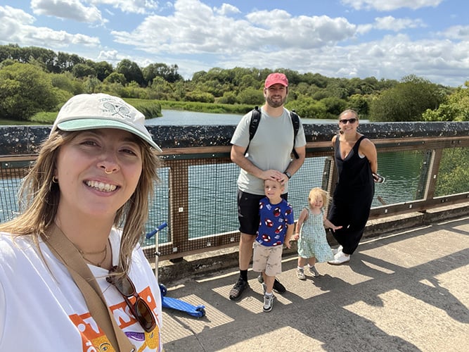 Group shot of three adults and two children on a bridge, with a lake surrounded by trees and cloudy blue sky behind.
