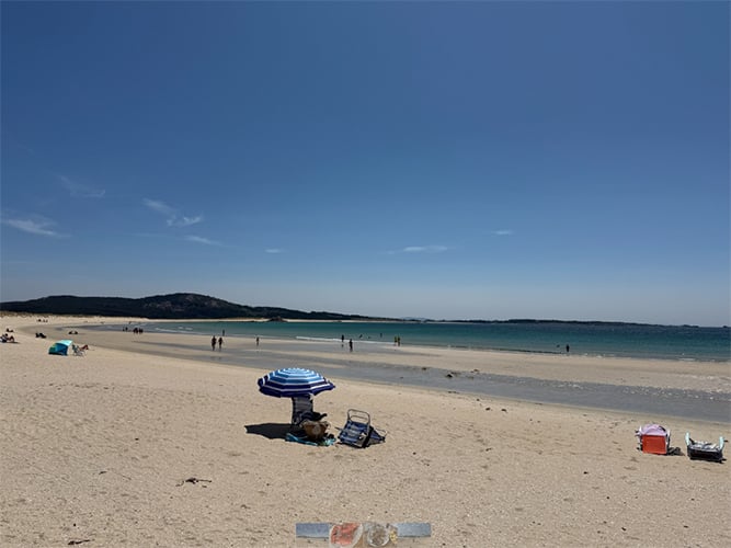 A sunny sandy beach with a blue and white striped umbrella shading a person, scattered beach chairs, calm ocean, and distant hills under a clear blue sky.