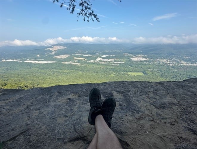 Person’s legs and black shoes resting on a rocky ledge overlooking a vast green valley with distant mountains under a partly cloudy blue sky.