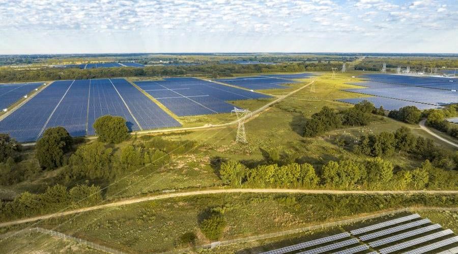Aerial view of a large solar farm with extensive rows of solar panels interspersed with green fields, trees, and power transmission towers under a partly cloudy sky.