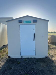 A small, white, prefabricated tornado shelter with a closed door, located outdoors on a gravel surface, with a sign above the door that reads "Pro Storm Shelters."