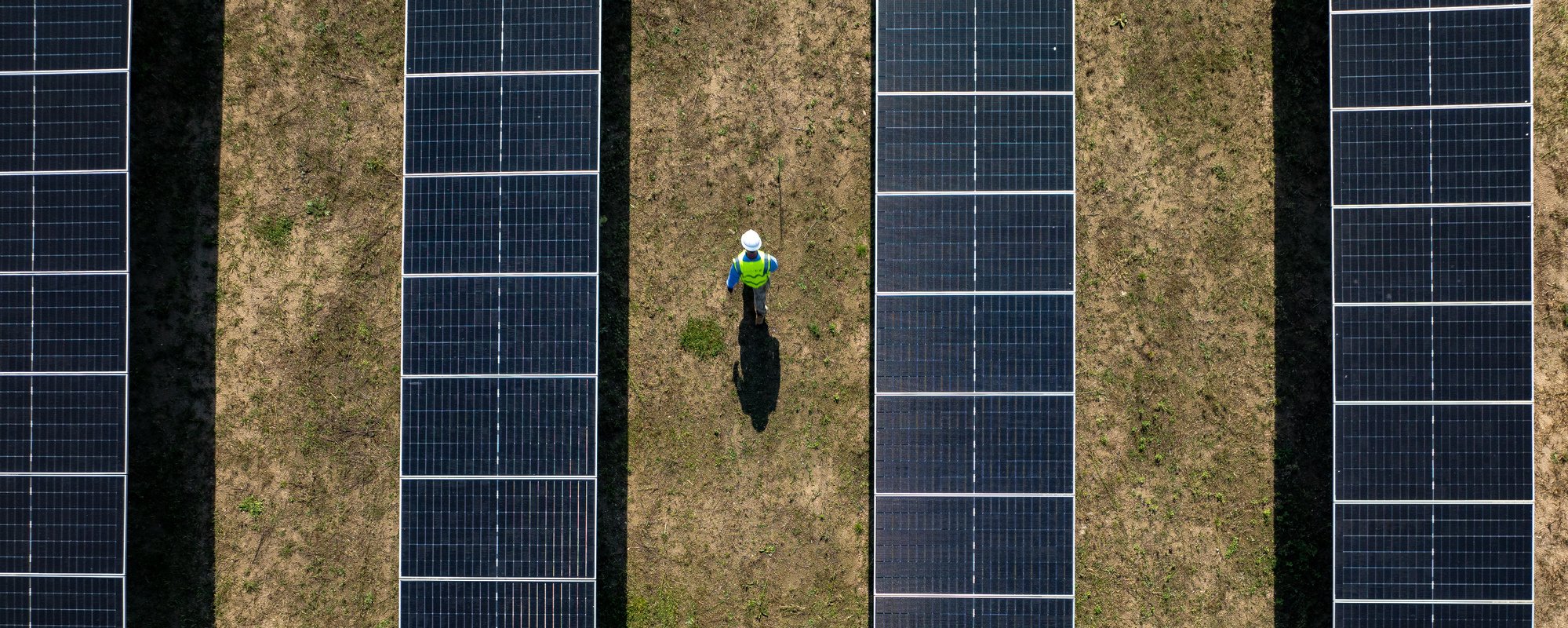 Aerial view of a worker wearing a high-visibility vest and hard hat walking between rows of solar panels.