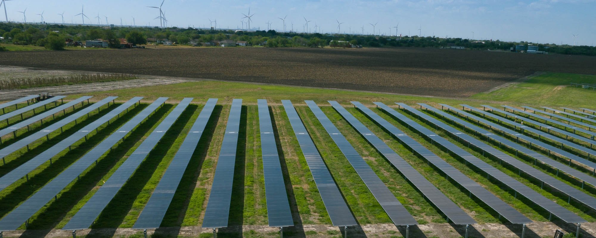 Rows of solar panels installed on green grassy land with a large ploughed field and a line of wind turbines visible in the distance under a clear blue sky.