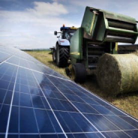 A tractor baling hay in a field alongside a solar farm, showcasing the concept of agrisolar land use where agriculture and renewable energy coexist.