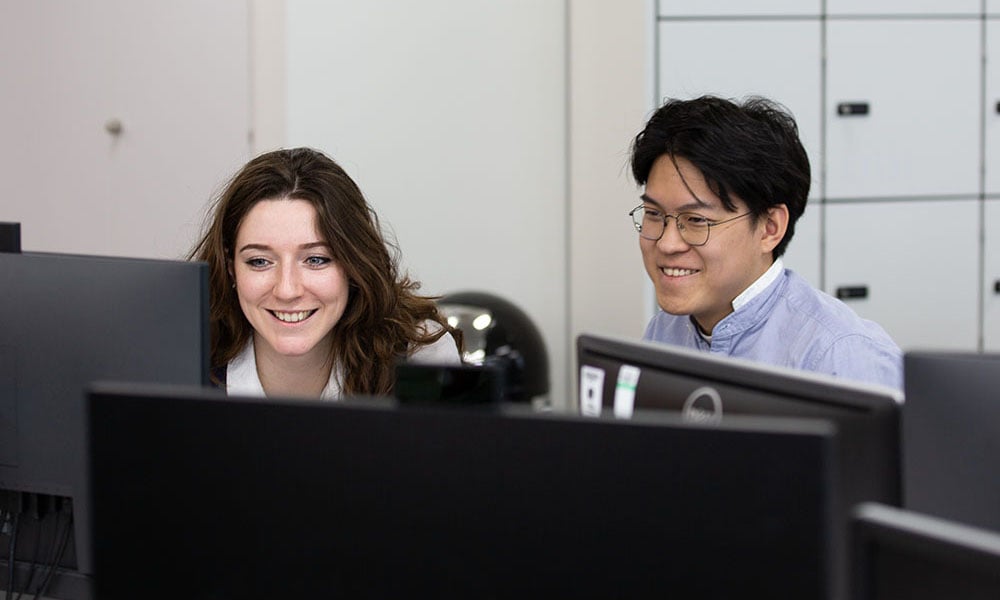 Two people, a female and male, sat at a desk with computer screens with smiling faces