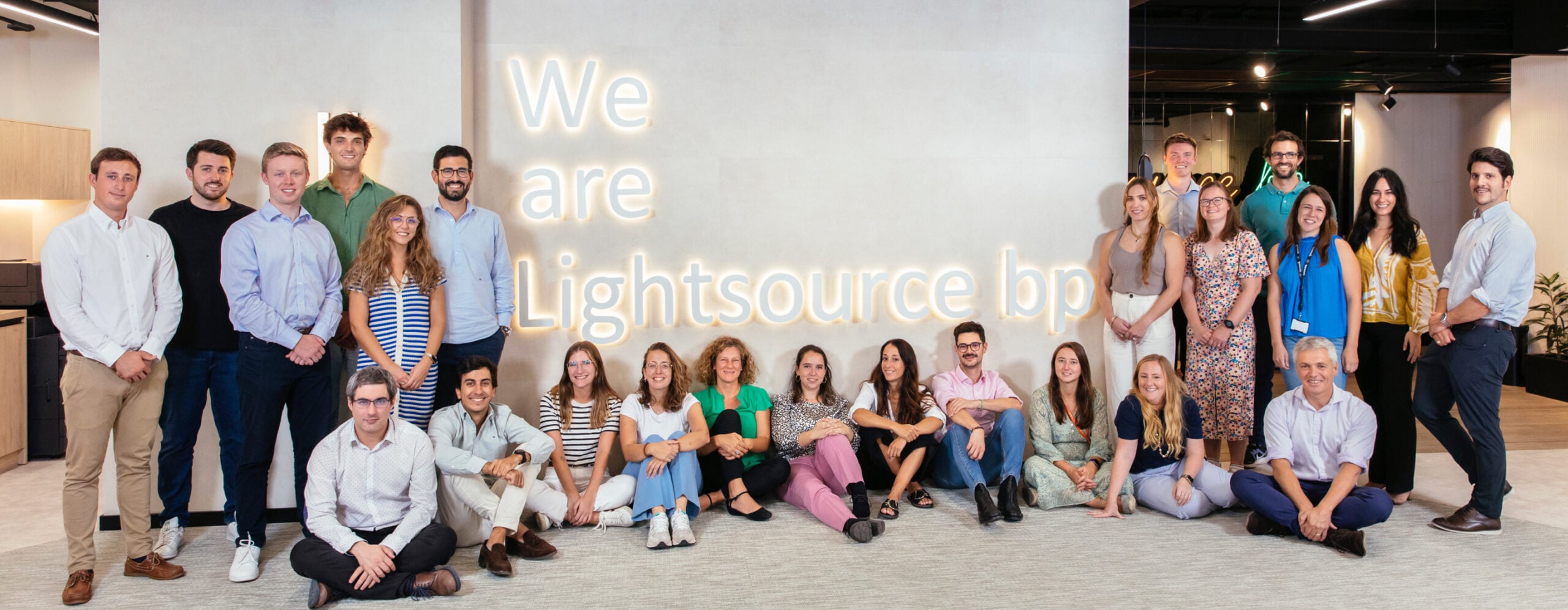 約26人的團體合影，部分人站立，部分人坐在地板上，背景是一面寫有「We are Lightsource bp」發光字樣的牆。