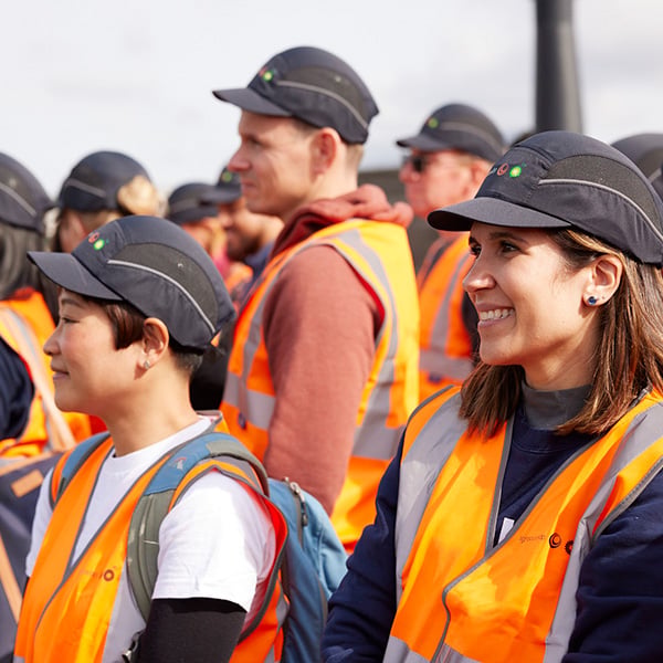 A group of people in orange safety vests and black caps are standing outdoors, attentively facing forward.