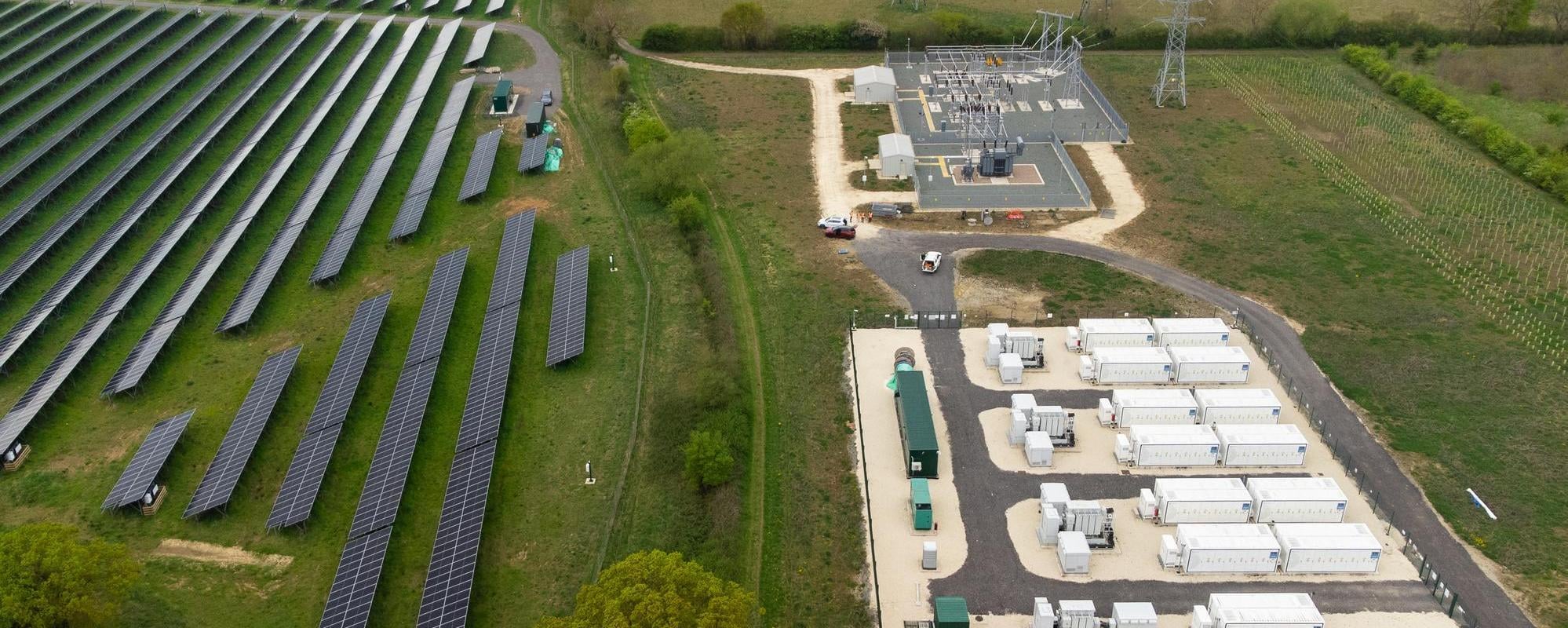 Aerial view of a renewable energy project with photovoltaic panels, battery storage containers, and electrical infrastructure integrated into agricultural land.