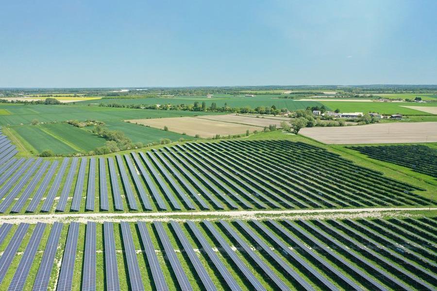 An aerial view of a large solar farm with rows of solar panels set among green fields and farmland under a clear blue sky.